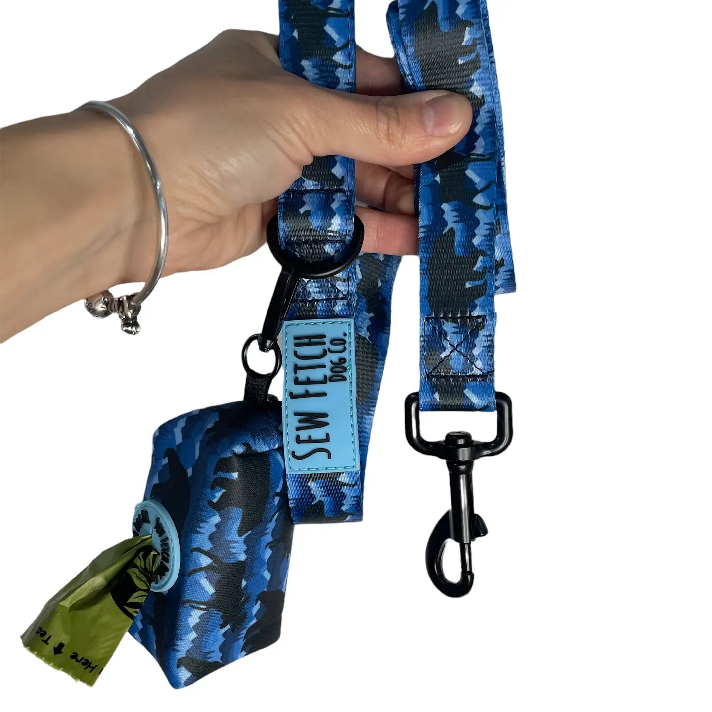 A hand holding a vibrant blue camo dog leash featuring a dark grizzly bear pattern, complete with a matching poop bag dispenser, matte black hardware, and a light blue rubber "Sew Fetch Dog Co." logo label.