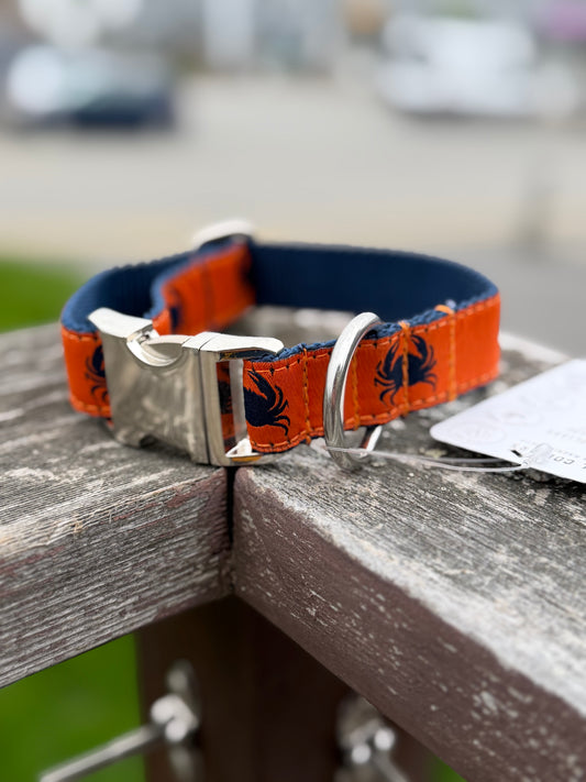 Close-up of a 1-inch wide adjustable dog collar resting on a wooden railing. It features an orange ribbon with a navy blue crab pattern sewn onto a navy backing, showcasing the durable, non-rusting metal quick-release clasp and metal D-ring for leash attachment.
