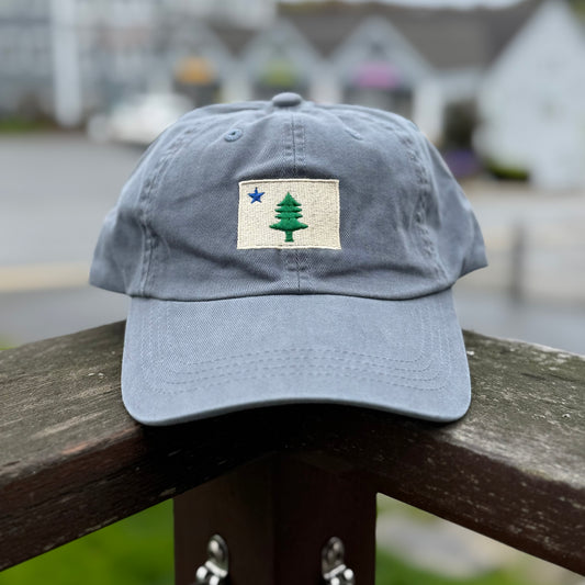 Slate blue Belted Cow dad hat with embroidered original Maine 1820 flag patch featuring a green pine tree and blue star.