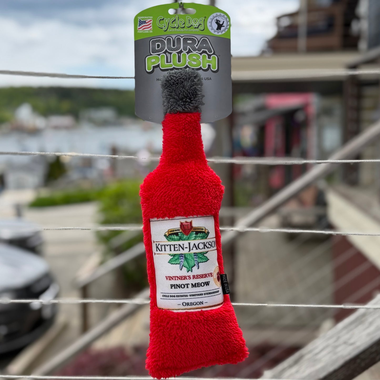 Red plush cat toy resembling a bottle with a label on a metal railing, against an outdoor background.