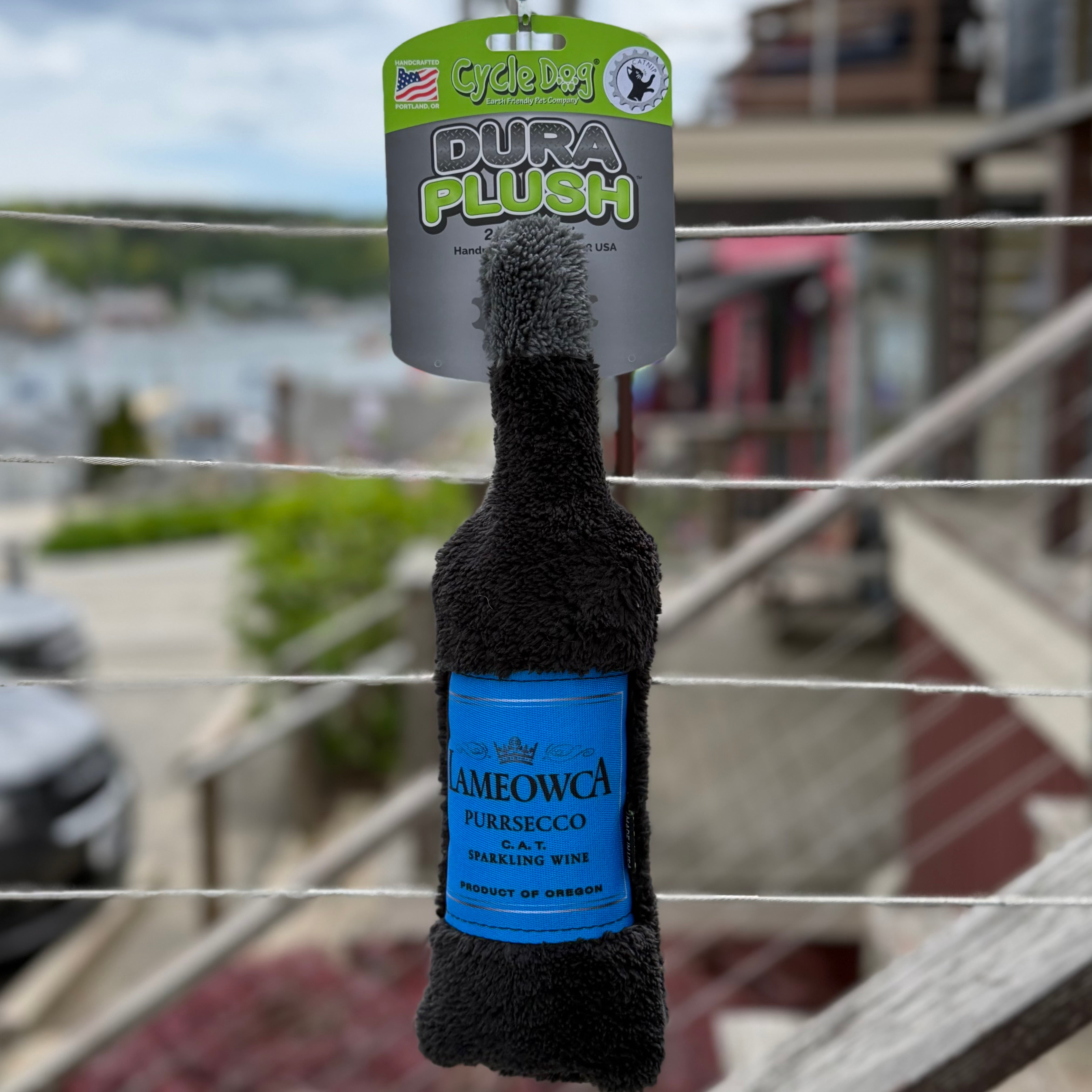 Wine bottle shaped cat toy with "Lameowca Purrsecco" text hanging with a blurry background.