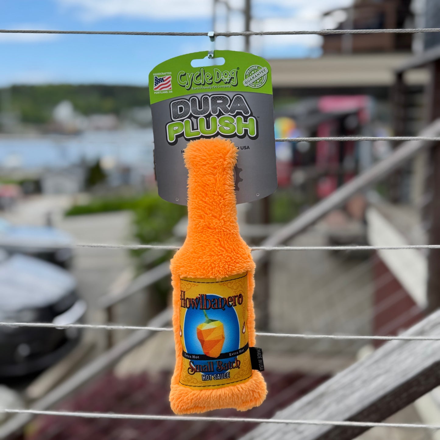 Orange bottle shaped plush dog toy with 'Howlbanero' branding on a metal railing with a blurred background